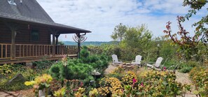 A wooden cabin with a deck, surrounded by a garden with various plants and flowers, and two Adirondack chairs. - Entire 70 acre private Ozark Mountain Retreat. Sleeping room for TEN guests! (Pelsor)