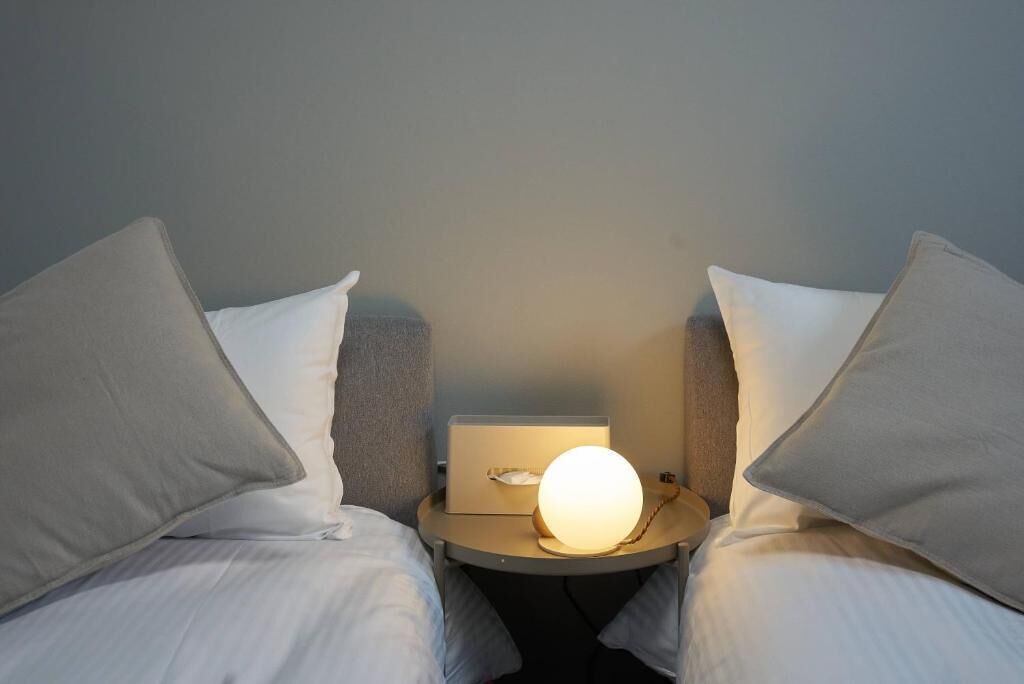 Una cama con dos plazas, ropa de cama blanca, una mesita de noche con lámpara y un mueble con caja de pañuelos.