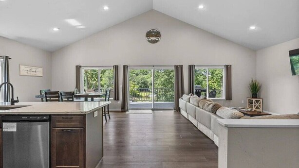 A spacious kitchen with a dishwasher, a dining area with chairs, a large window, and a living area with a sofa and a TV.