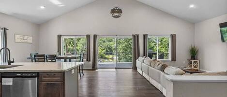 A spacious kitchen with a dishwasher, a dining area with chairs, a large window, and a living area with a sofa and a TV.