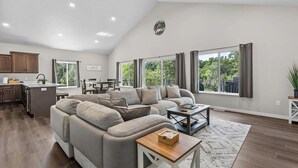 A spacious living area with a large gray sectional sofa, a dining table, and a kitchen area with wooden cabinets and a window overlooking greenery.
