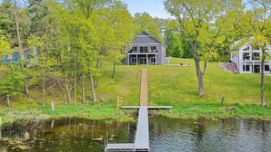 A modern house with a dock extending into a lake, surrounded by lush greenery and trees.