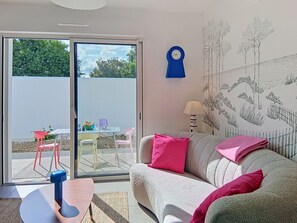 Ein Zimmer mit einer Couch, einem Tisch, einer Lampe, einer Wanduhr und einer Glastür, die zu einem Außenbereich mit Stühlen und einem Tisch führt.