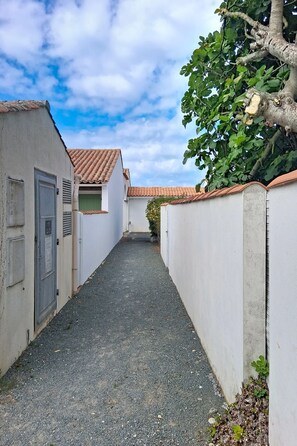 A narrow alleyway between buildings with a gravel path, a tree on the right, and a clear sky. - Les Dunes by Interhome (Ile d'Oléron)