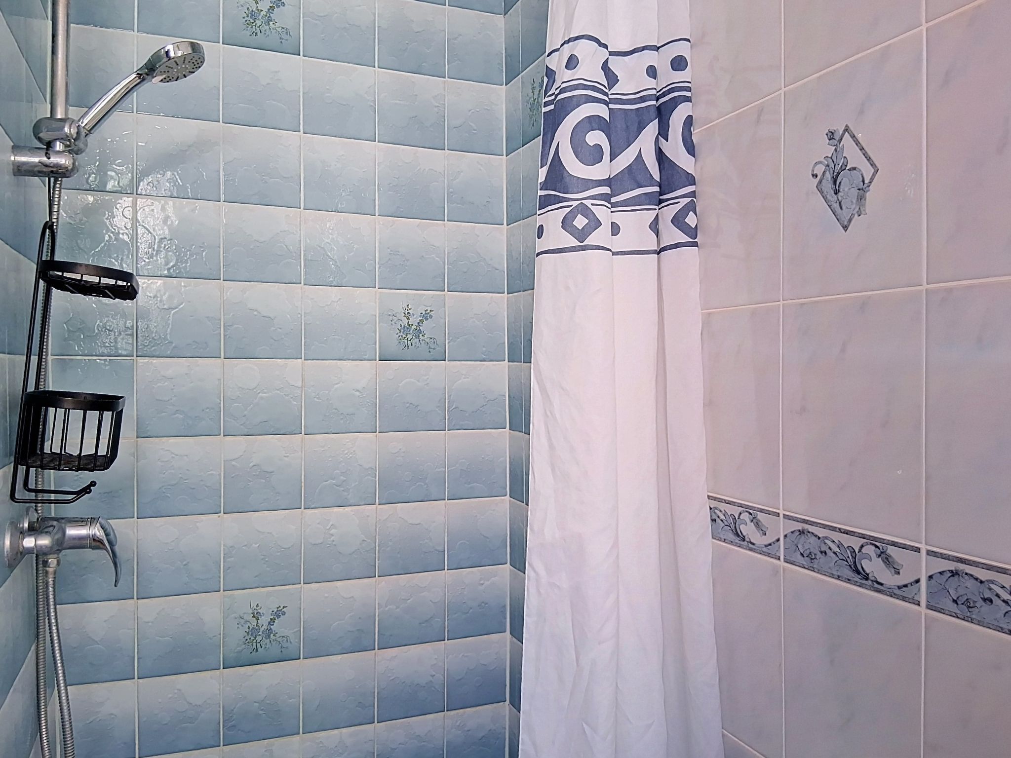 A bathroom with blue patterned tiles, a shower with a handheld showerhead, a black metal basket, and a white shower curtain with a decorative border.