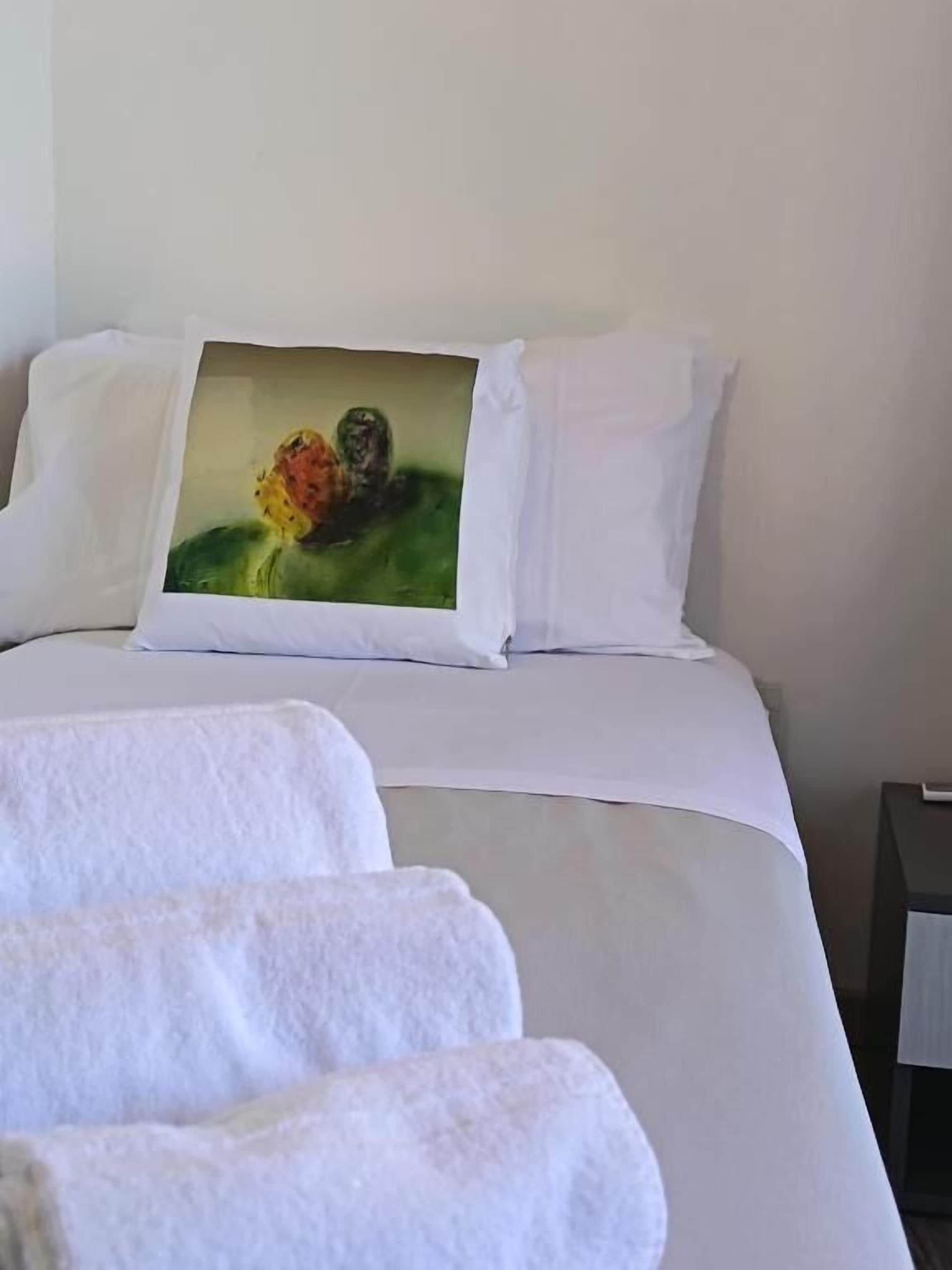 Una cama con sábanas blancas, un cuadro en la cabecera y una toalla blanca doblada sobre la cama.
