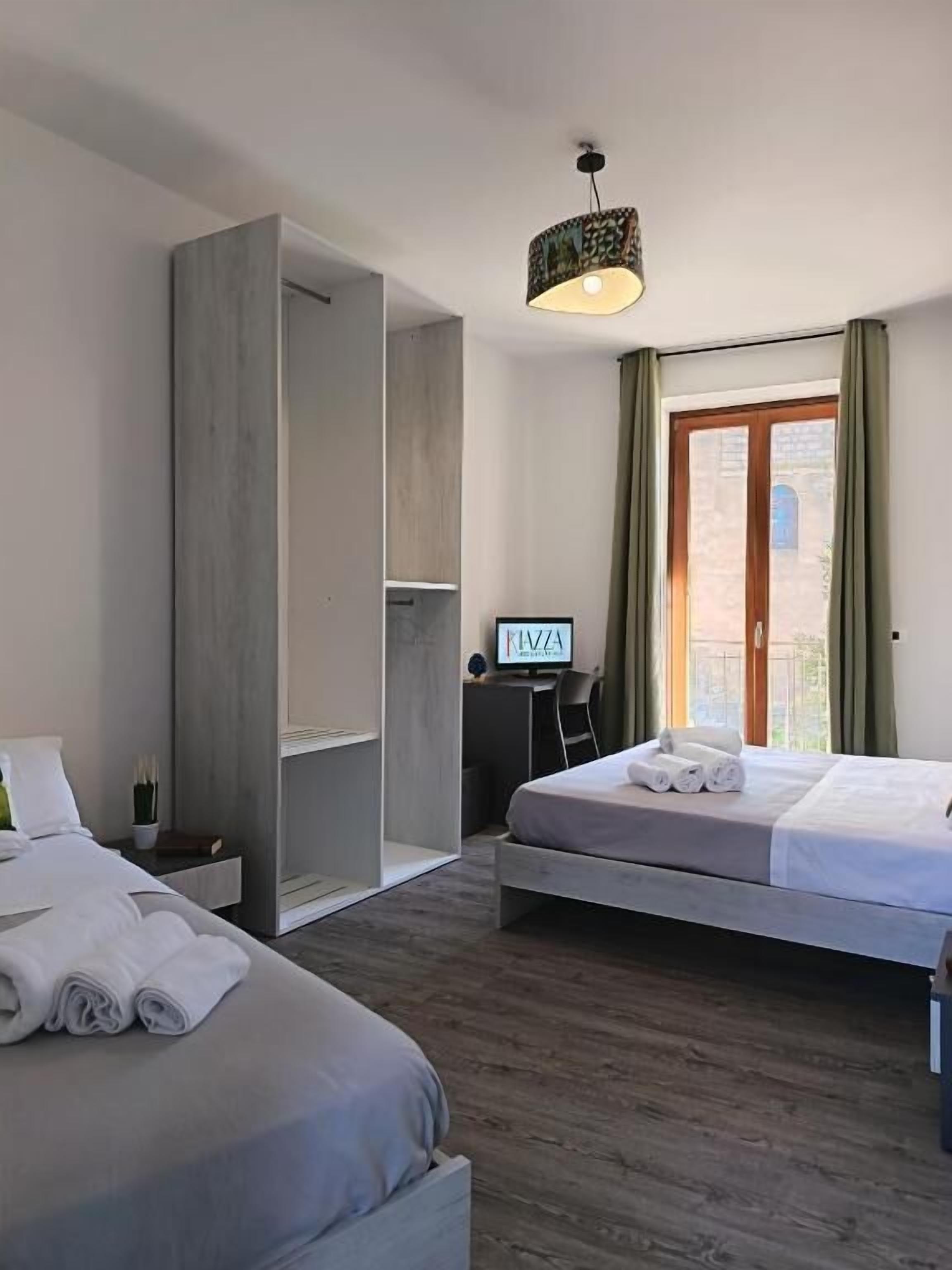 A hotel room with two beds, a desk, a chair, a hanging light fixture, and a view of the outdoors.