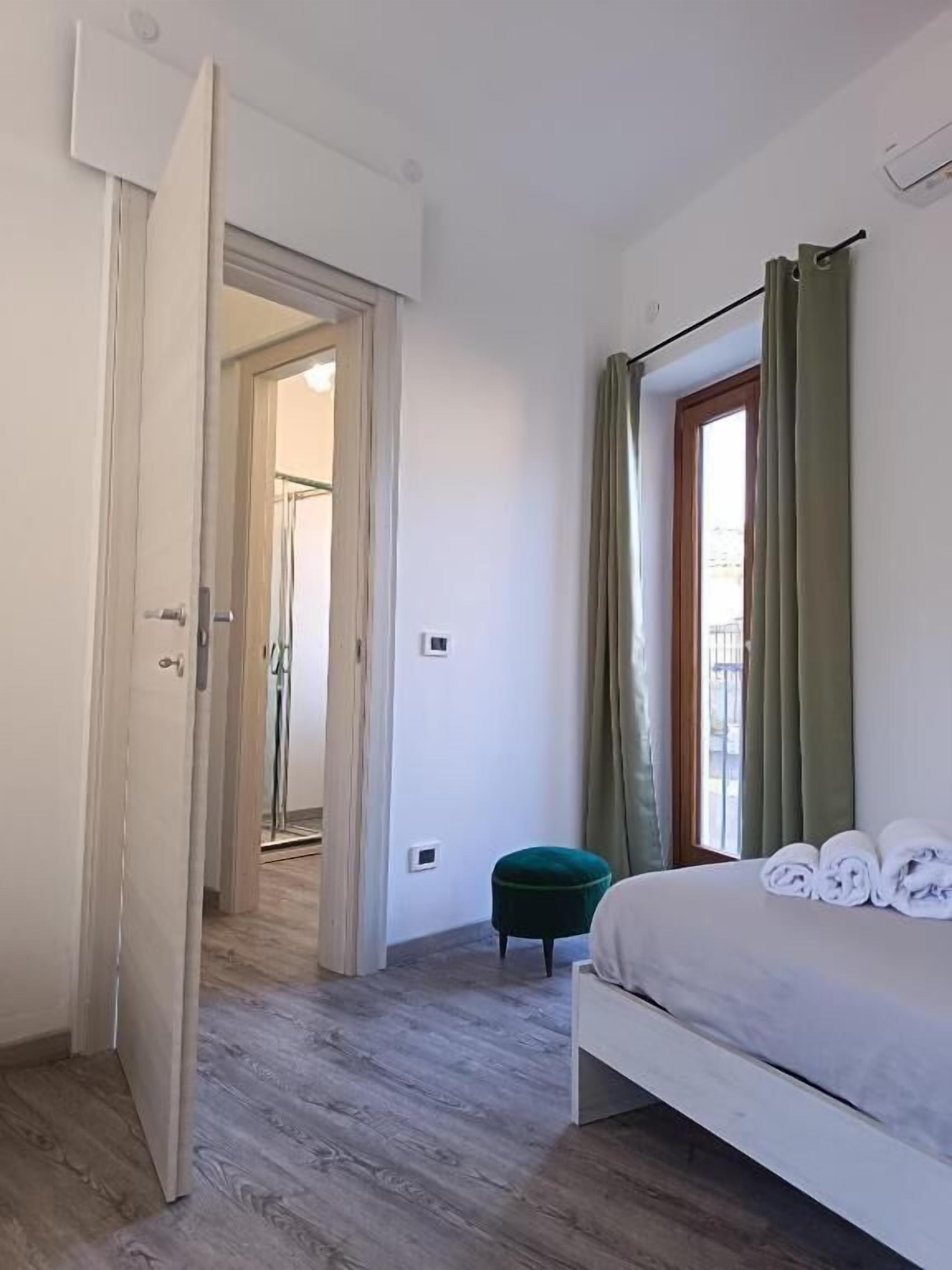 A bedroom with a bed, a green ottoman, a door leading to another room, and a window with curtains.