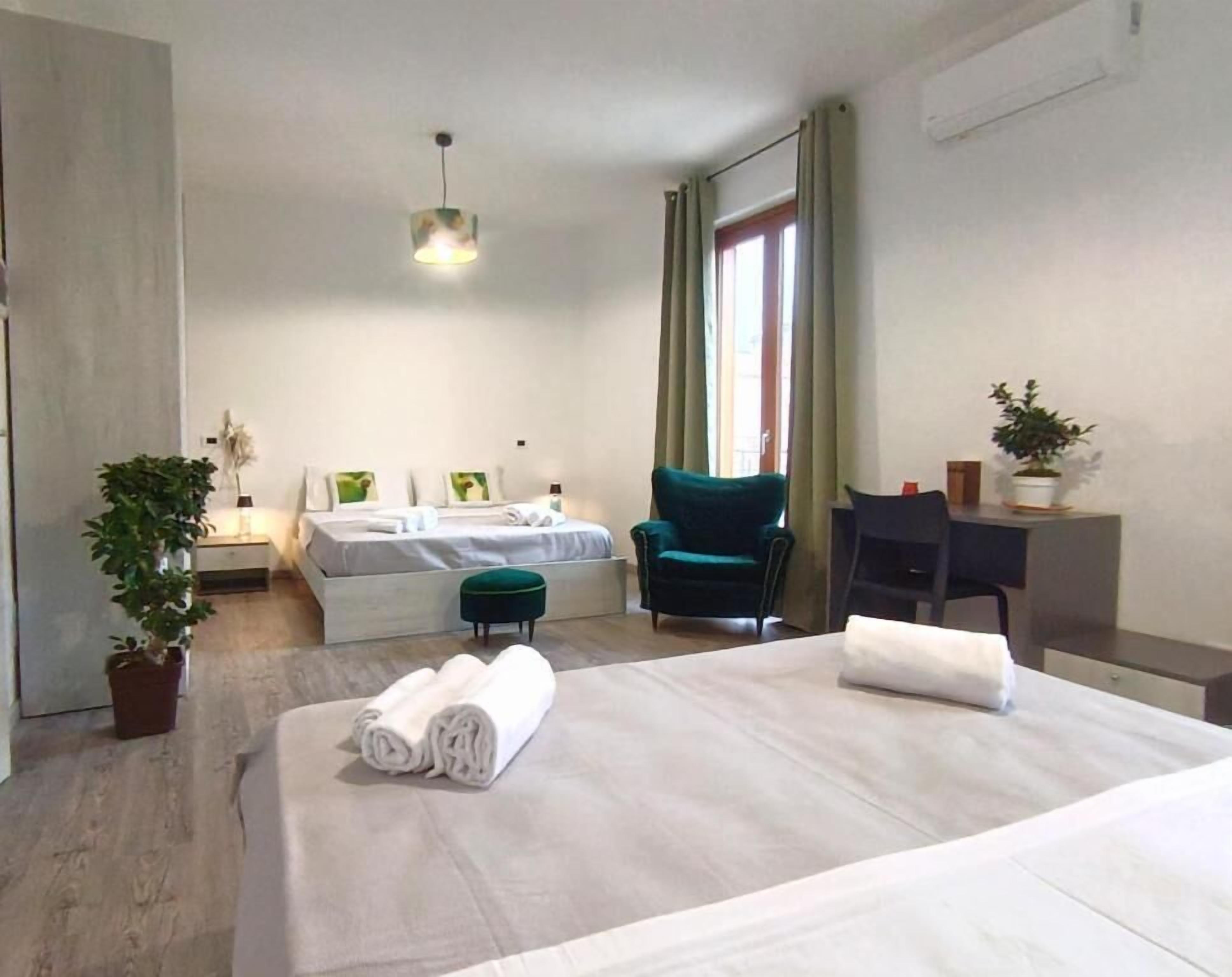 Habitación de hotel con cama, escritorio, silla, mesita, planta en maceta y ventana con cortinas.