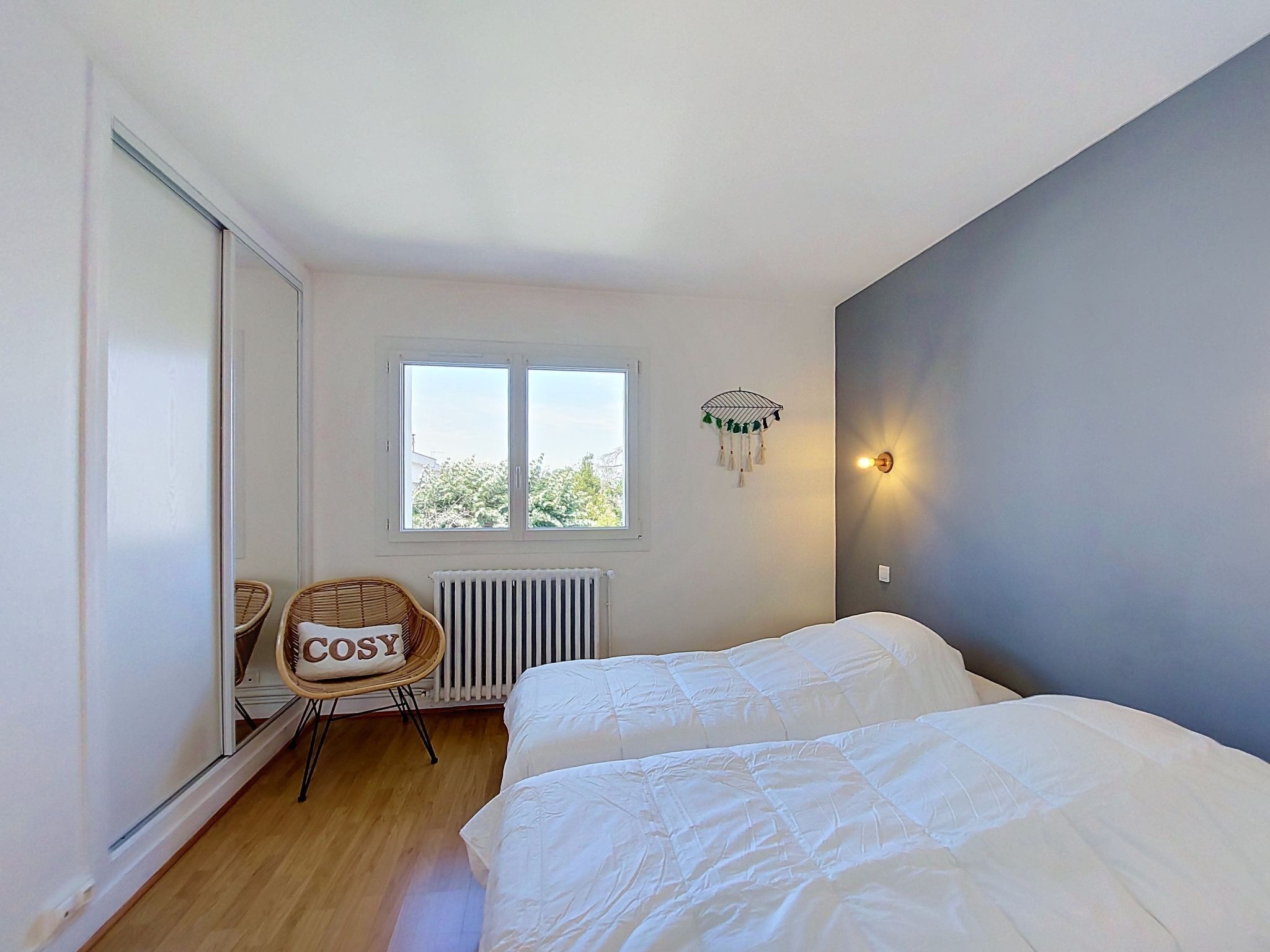 A bedroom with two beds, a radiator, a wicker chair, a window, and a wall-mounted light fixture.