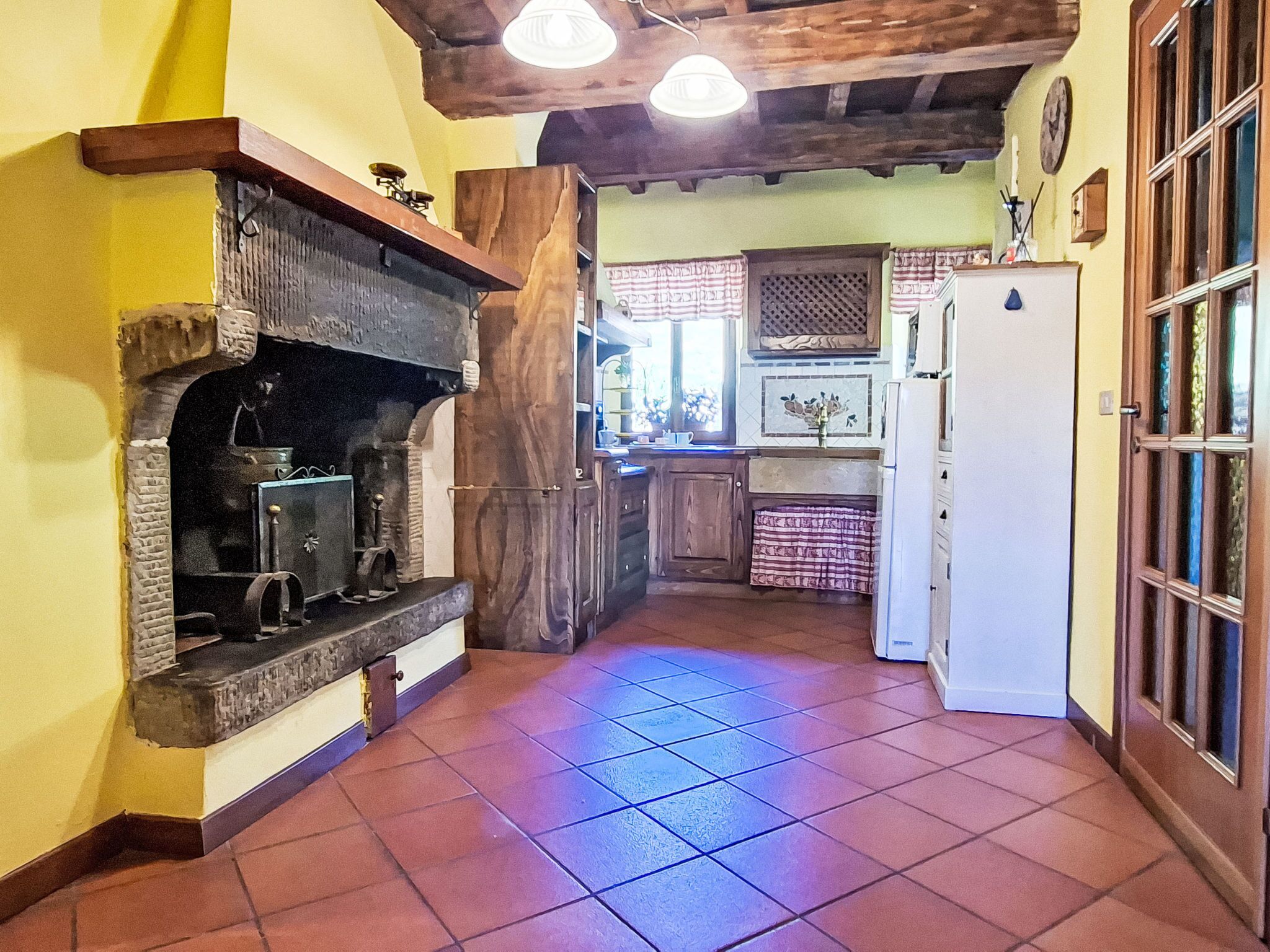 A kitchen with a stone fireplace, wooden beams, a tiled floor, and a refrigerator.