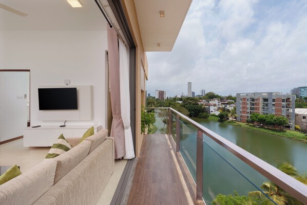 Una sala de estar moderna con un televisor de pantalla plana, un sofá y un balcón con vistas a un río y un paisaje urbano al fondo.