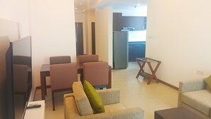 A living room with a TV, sofa, and chairs. There is a kitchen in the background.