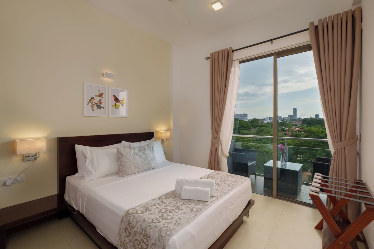 Habitación de hotel con cama, dos lámparas de pared, ventana con cortinas y vista a la ciudad.