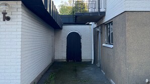 Une ruelle étroite, avec un portail noir, des murs en briques blanches et un petit espace aménagé en jardin.