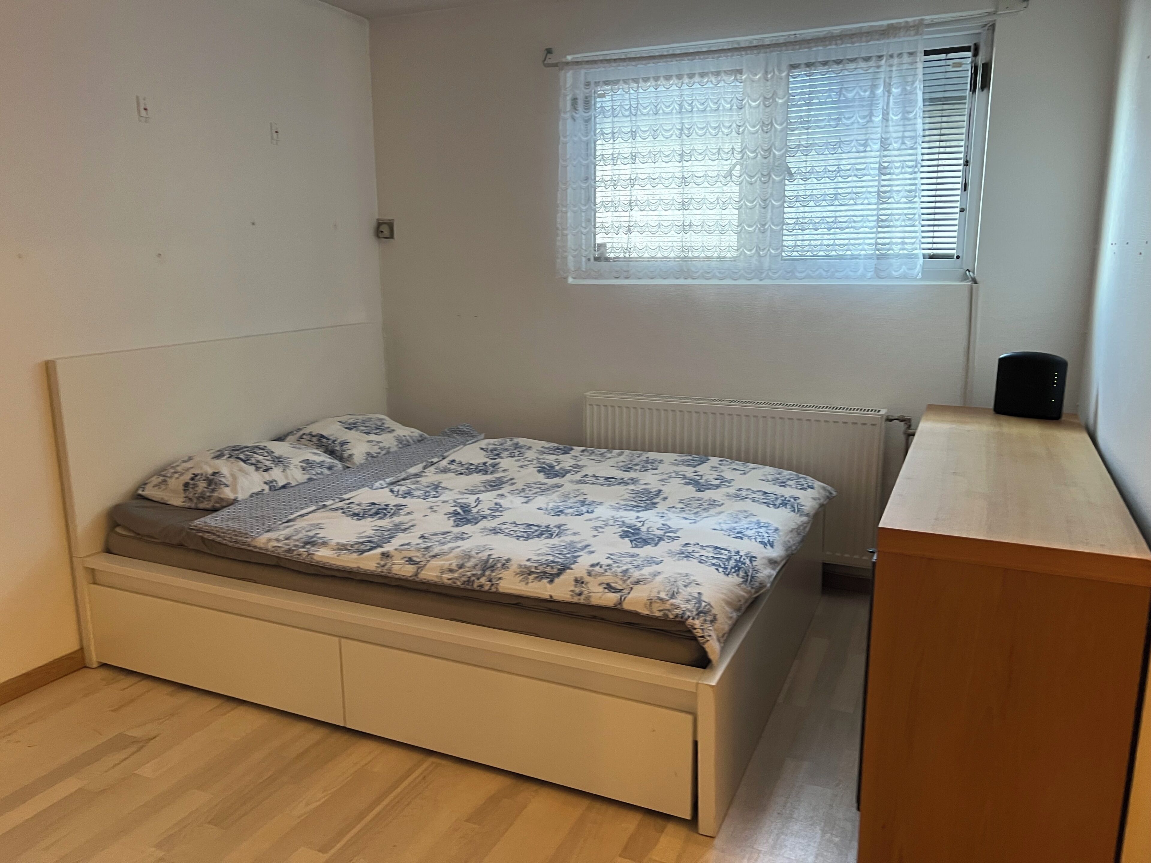 A single bed with a patterned bedspread, a radiator, a window with blinds, and a small wooden desk.