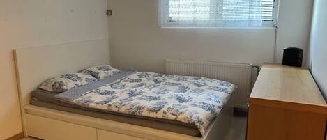 A single bed with a patterned bedspread, a radiator, a window with blinds, and a small wooden desk.
