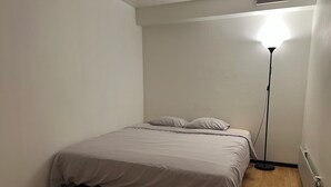 Un lit simple avec une couvre-lit grise, dans une pièce au plancher en bois et dotée d’un lampadaire.