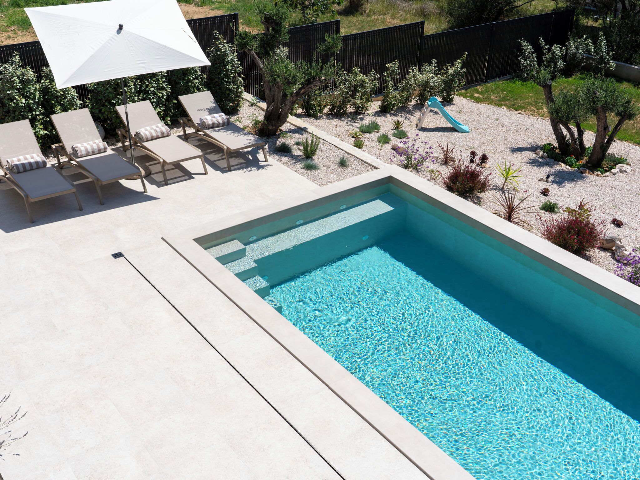 A rectangular swimming pool with clear blue water, surrounded by a concrete deck. There are lounge chairs and a white umbrella.
