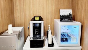 A room with a coffee machine, a black device with a pump, a white dispenser, and a refrigerator displaying beverages. - Gimhae H. Avenue (Gimhae)