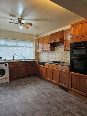 A kitchen with wooden cabinets, a built-in oven, a dishwasher, and a ceiling fan. - Big 3 Bedroom house 4 min walk from Chadwell Heath Station. (Dagenham)