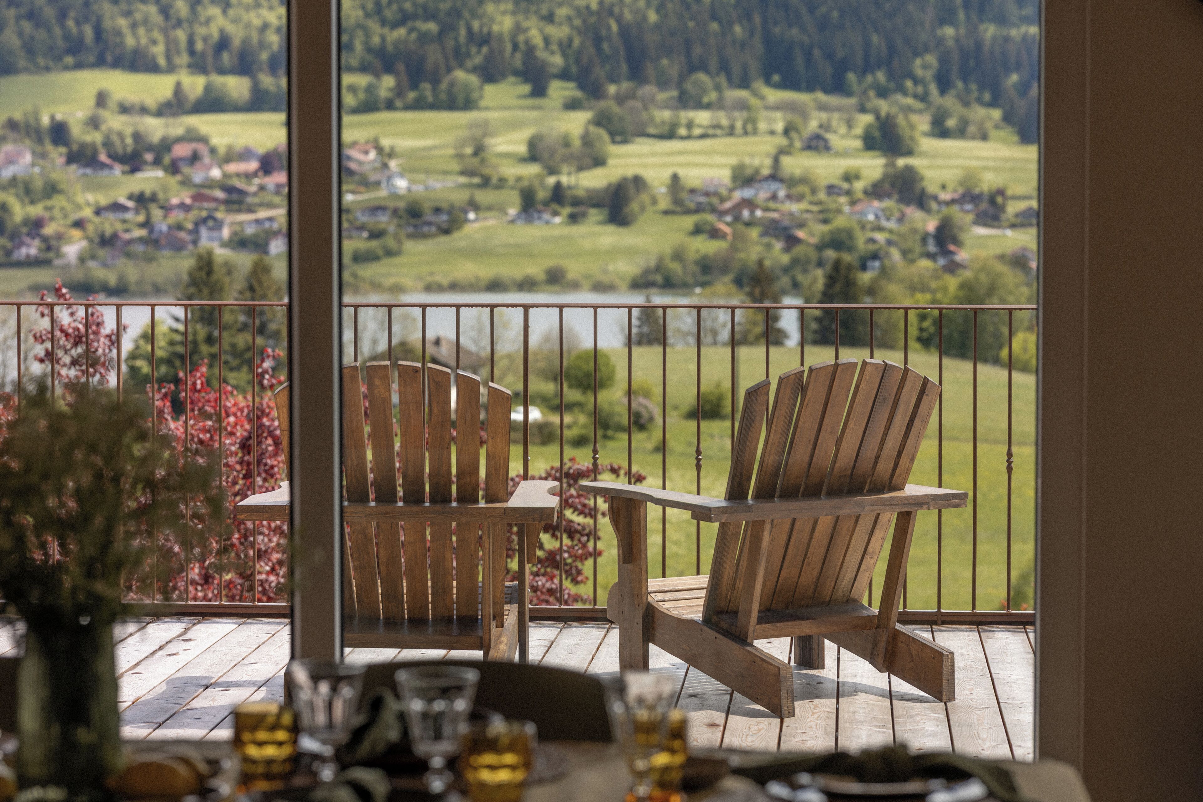 Ein Balkon mit zwei Holz-Adirondack-Stühlen und einem Tisch, der eine malerische Landschaft mit Feldern und Häusern überblickt.