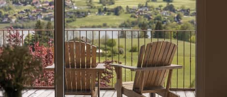 Een balkon met twee houten Adirondack-stoelen en een tafel, met uitzicht op een landschap met velden en huizen.