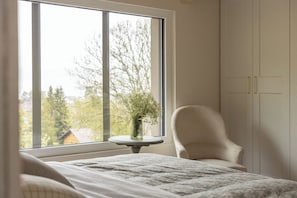 A bedroom with a large window, a chair, a bedside table with a vase, and a bed with a patterned bedspread. - Between the Lake and the Jura, Luxury Home—Sleeps 6—View of Lake St. Point: Luxury and Cozy Comfort (Les Grangettes)