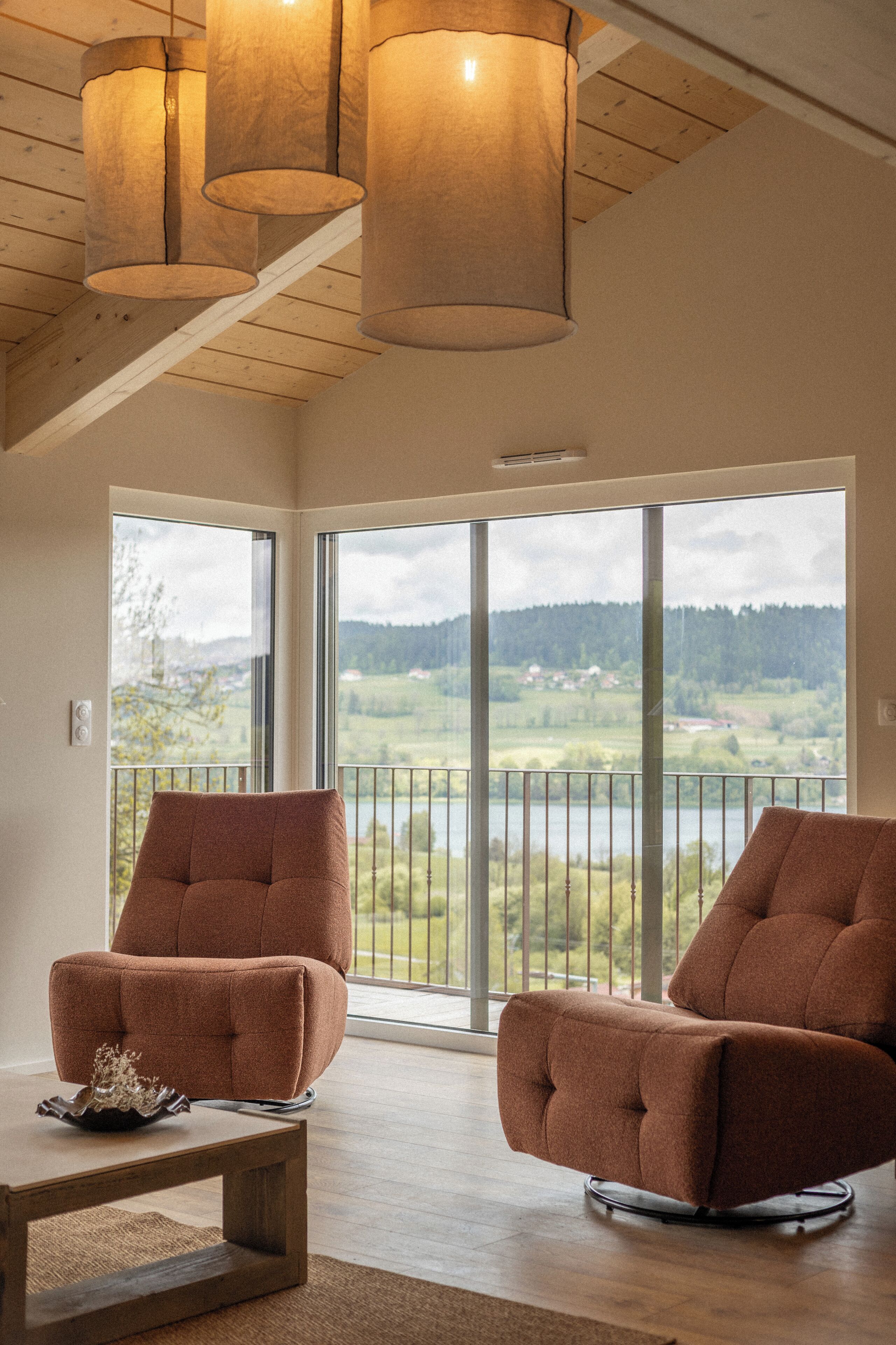 Ein geräumiges Zimmer mit zwei braunen Sesseln, einem Holztisch und großen Fenstern mit Blick auf eine Landschaft.