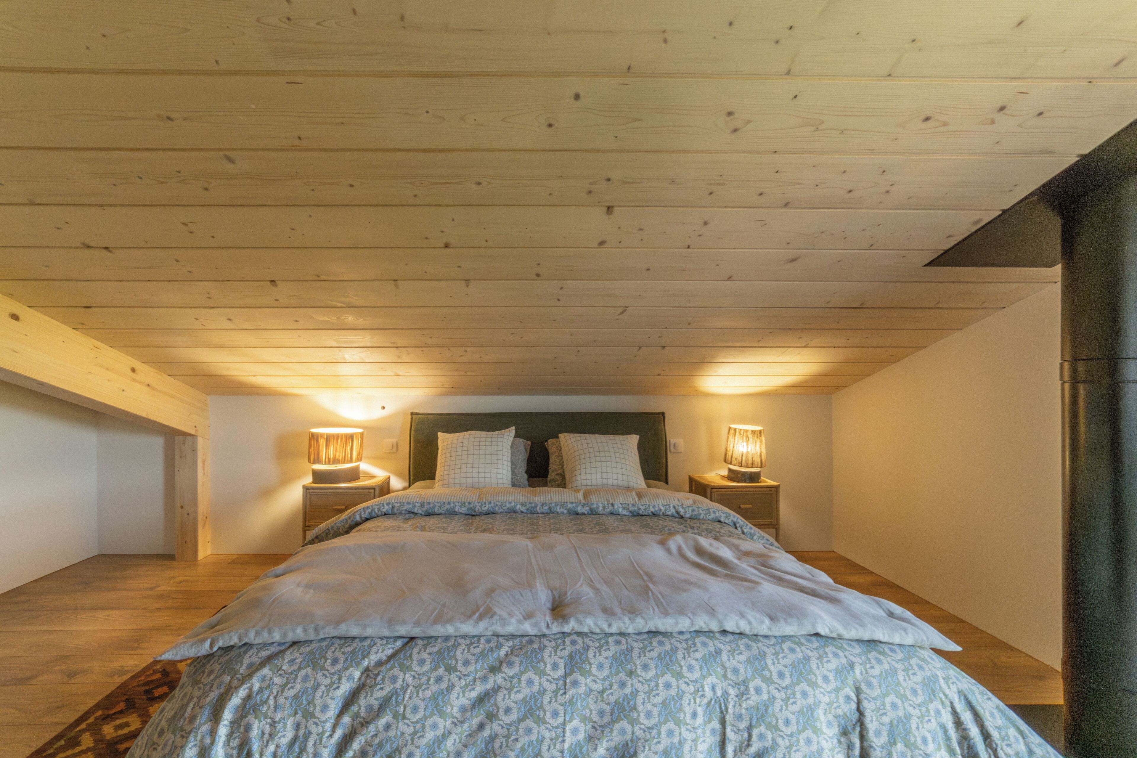 Un dormitorio con techo de madera, una cama con ropa de cama estampada, dos lámparas de noche y una mesita de noche.