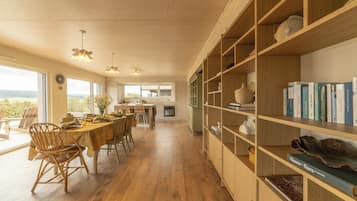 Een ruime eetruimte met een houten tafel en stoelen, een boekenplank vol boeken en uitzicht door grote ramen.
