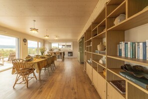 A spacious dining area with a wooden table and chairs, a bookshelf filled with books, and a view through large windows. - Between the Lake and the Jura, Luxury Home—Sleeps 6—View of Lake St. Point: Luxury and Cozy Comfort (Les Grangettes)