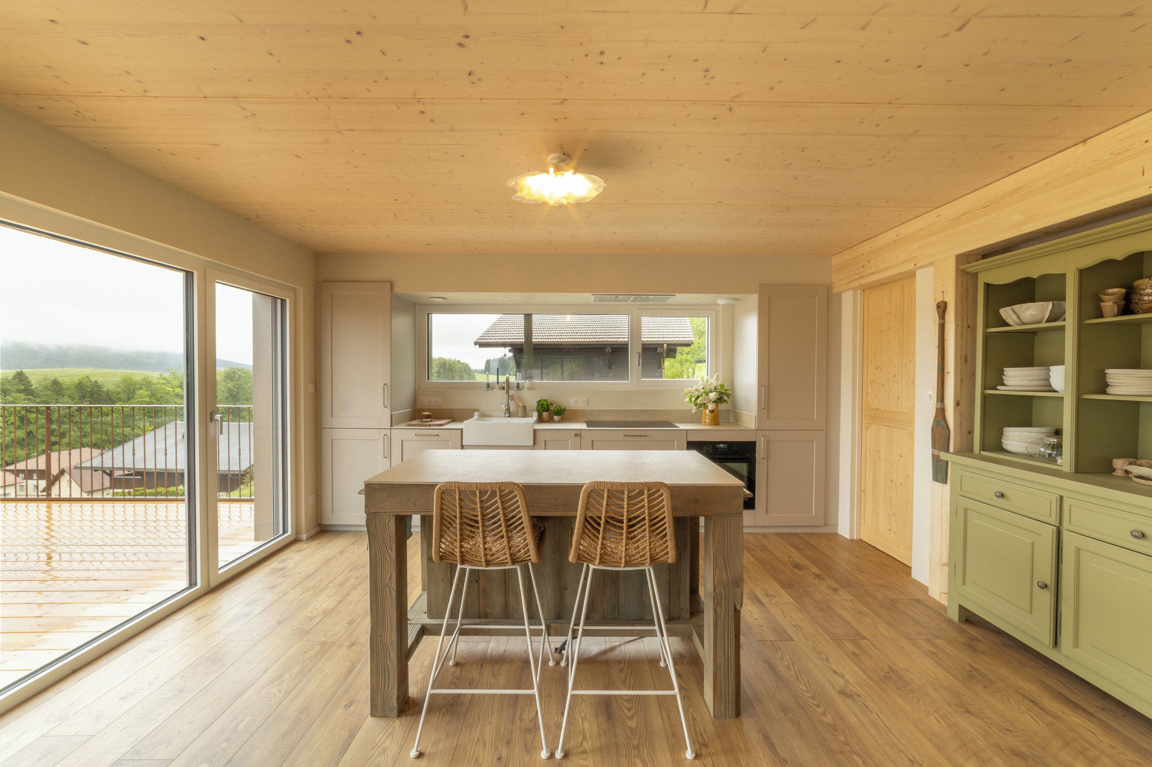 Eine moderne Küche mit Holztisch und -stühlen, Blick ins Freie und einem grünen Schrank mit Geschirr.