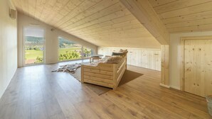 Une chambre spacieuse située dans les combles, avec un plafond en bois, une grande fenêtre donnant sur la campagne, un lit recouvert d’un plaid en fourrure et une petite table.