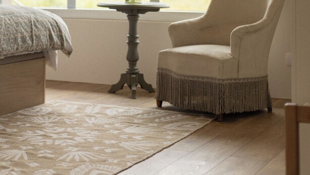 Une pièce avec une grande fenêtre, une chaise, une petite table sur laquelle se trouve un vase, et un lit recouvert d’une couvre-lit à motifs.