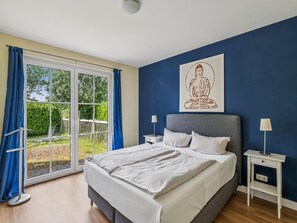A bedroom with a bed, a nightstand, a lamp, a window with curtains, and a wall with a framed picture. - Coastal Holiday Apartment near Kühlungsborn (Bastorf)