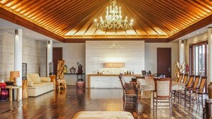 A spacious living area with a high wooden ceiling, a chandelier, a sofa, a dining table with chairs, and a fireplace.
