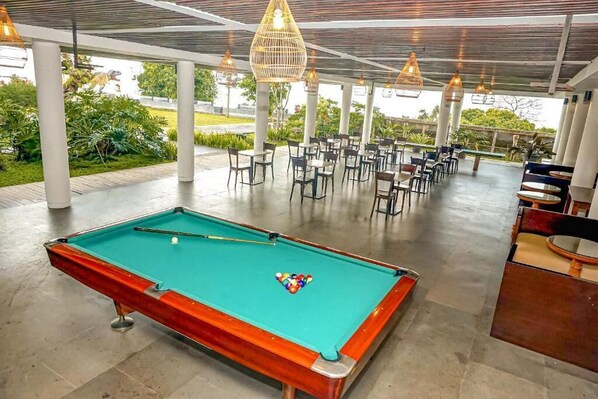 A pool table with a green felt surface, surrounded by chairs and tables in an indoor setting with a view of greenery outside.