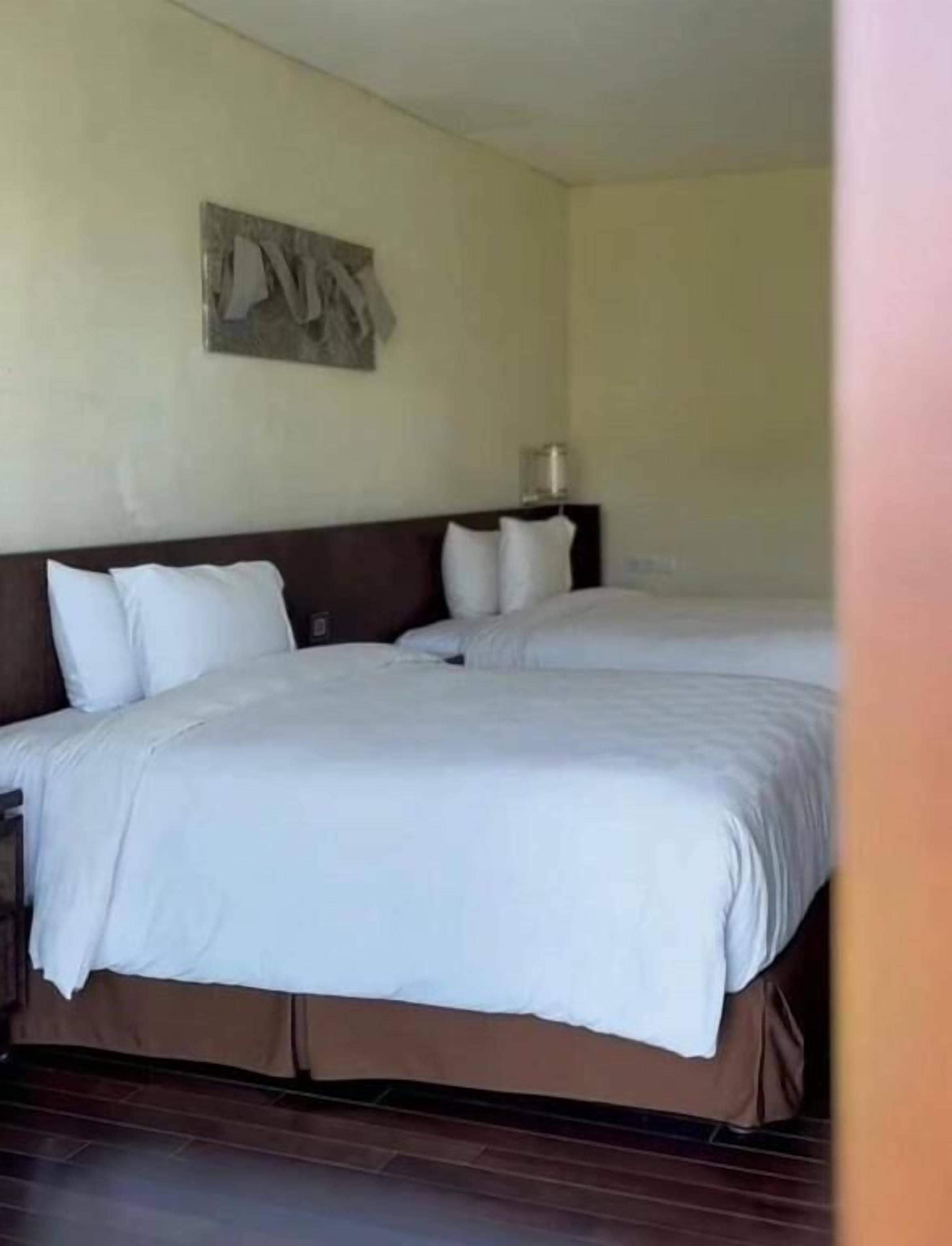 A hotel room with two beds, a headboard, a wall-mounted artwork, and a bedside table with a lamp.