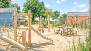 Un parque infantil con estructuras de madera, un tobogán, mesas de picnic y un edificio con tejado de baldosas.