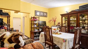 Un coin repas avec une table dressée pour quatre personnes, entourée de chaises en bois. Un ours en peluche est posé sur le sol, et une bibliothèque remplie de divers objets se trouve à gauche.