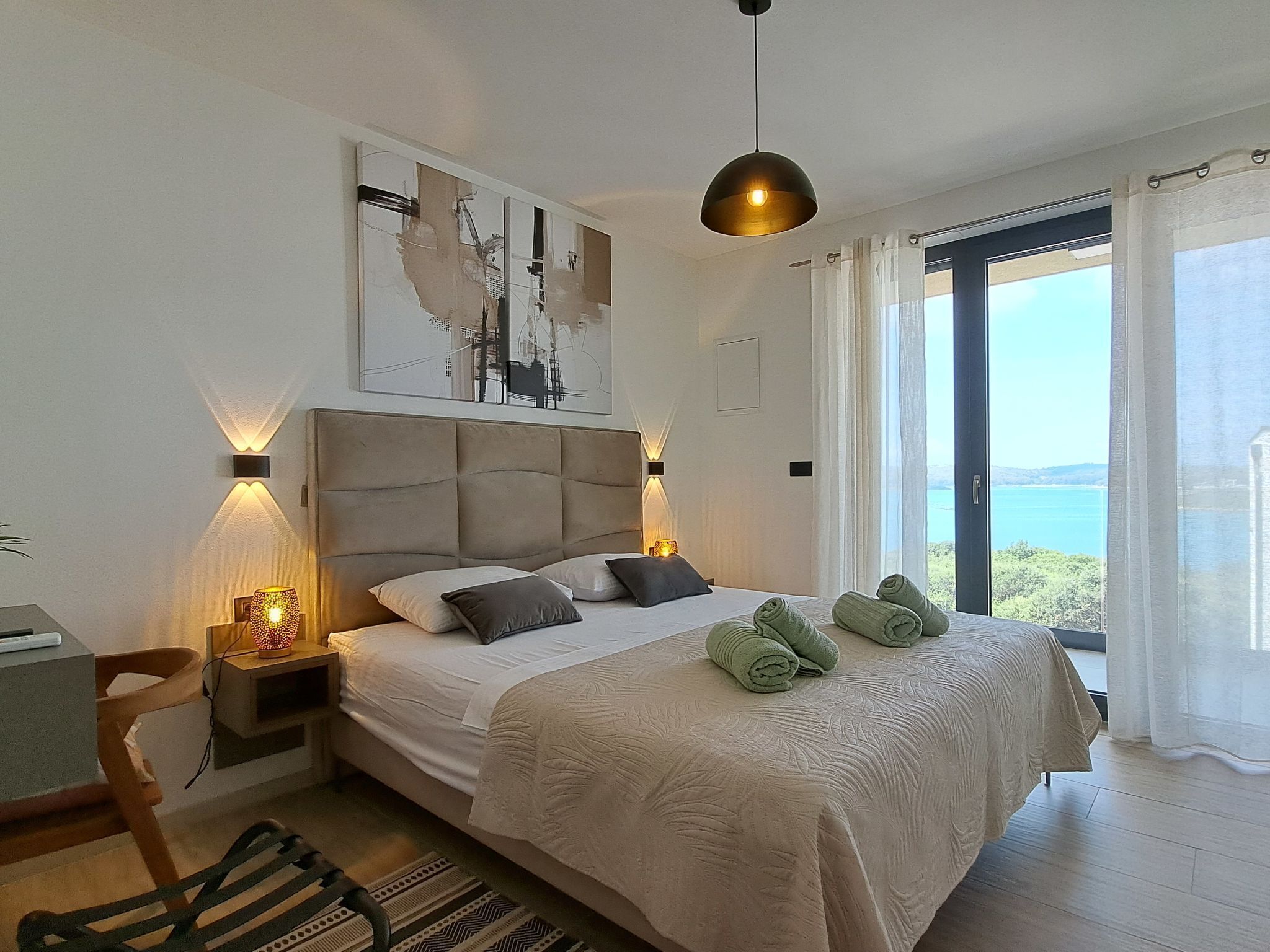 A bedroom with a large bed, two bedside lamps, a view of the sea, and a painting on the wall.