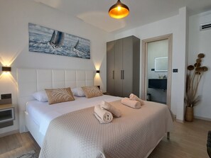 Ein modernes Schlafzimmer mit einem großen Bett, einem Gemälde von Segelbooten, einem Kleiderschrank und einem durch eine offene Tür einsehbaren Badezimmer.