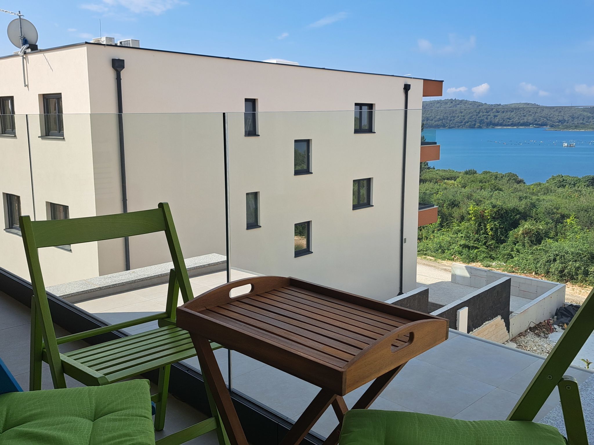 Un balcon avec une table en bois et des chaises vertes, donnant sur un bâtiment moderne et une étendue d’eau.