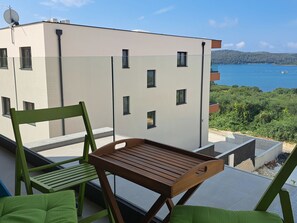 Ein Balkon mit einem Holztisch und grünen Stühlen, mit Blick auf ein modernes Gebäude und ein Gewässer.