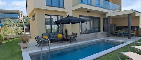 A modern two-story house with a swimming pool, outdoor furniture, and a covered patio area.