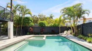 Et rektangulært pool med et lille spa, omgivet af et træhegn, palmer og en overdækket patio.