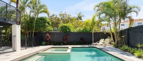 A rectangular swimming pool with a small hot tub, surrounded by a wooden fence, palm trees, and a covered patio area.