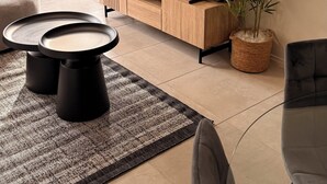 Ett modernt vardagsrum med en platt-TV, en grå soffa, ett träbord, ett svart soffbord, en mönstrad matta, en krukväxt och en matplats med ett glasbord och stolar.