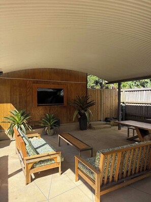 An outdoor patio with wooden furniture, a TV mounted on the wall, and potted plants. - Barefoot@Belongil (Byron Bay)